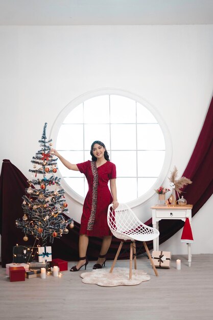 Portrait of a pretty young girl wearing a red gown smiling at the camera standing in decorated Christmas living room indoors