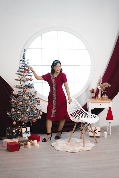 Portrait of a pretty young girl wearing a red gown smiling at the camera standing in decorated Christmas living room indoors