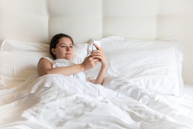 Portrait pretty young girl with smart phone on bed in the morning