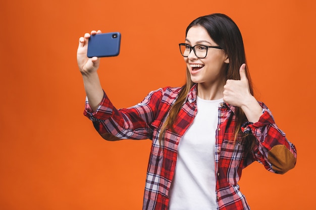 Portrait of pretty young happy woman making selfie on smartphone, isolated against orange background. Thumbs up.