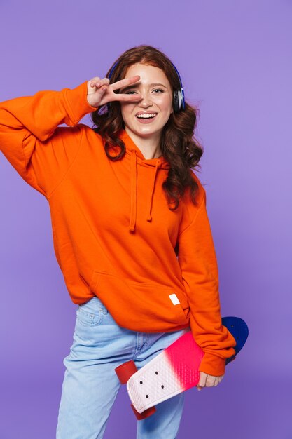 Portrait of a pretty young redheaded woman standing over violet, holding skateboard, listening to music with headphones