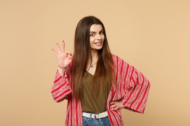 Portrait of pretty young woman in casual clothes looking camera, showing OK gesture isolated on pastel beige wall background in studio. People sincere emotions, lifestyle concept. Mock up copy space.