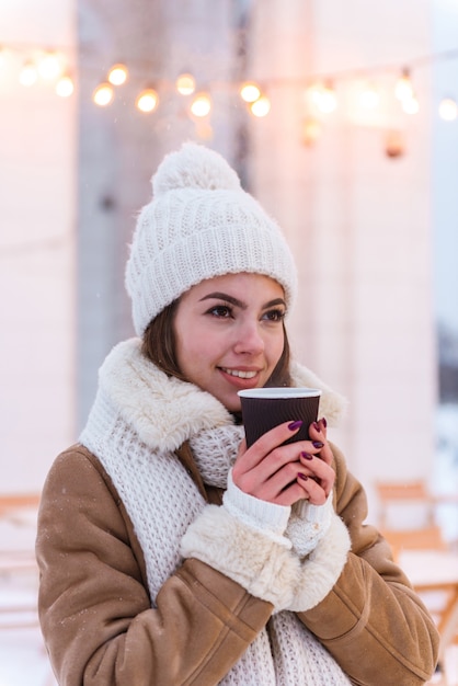 portrait of a pretty young woman in hat and scarf walking outdoors in winter snow drinking coffee.