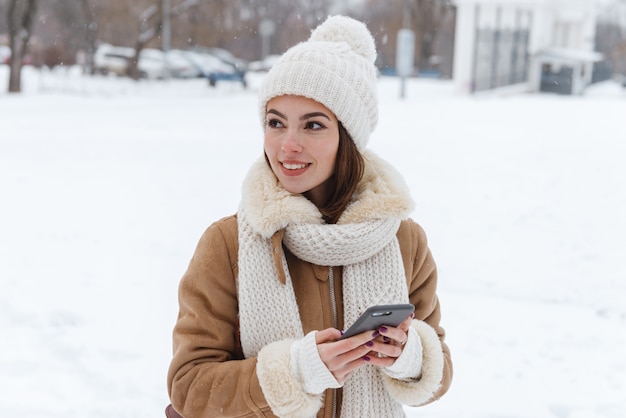 portrait of a pretty young woman in hat and scarf walking outdoors in winter snow using mobile phone.