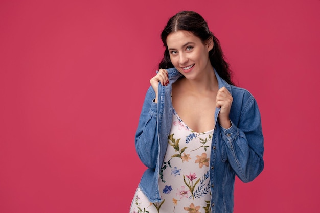 Portrait of a pretty young woman in a light dress and blue shirt standing on pink background in studio. People sincere emotions.