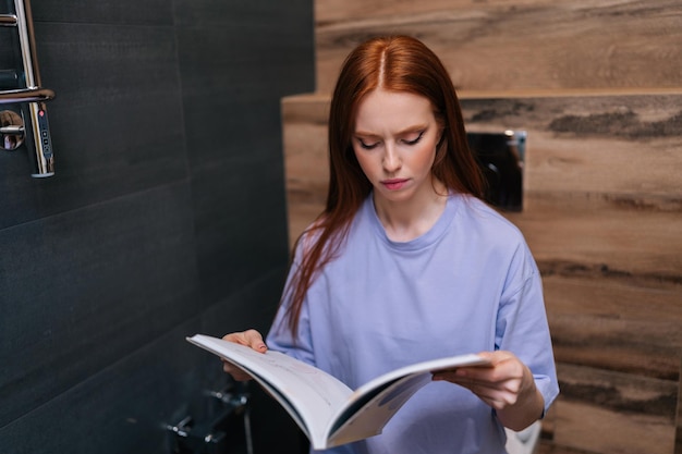 Portrait of pretty young woman reading fashion magazine sitting on toilet bowl at home restroom with modern interior Redhead female reading paper book