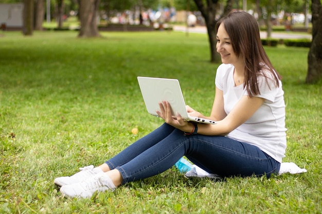 Portrait of pretty young woman sitting on green grass in park with laptop. Happy woman, student, fre