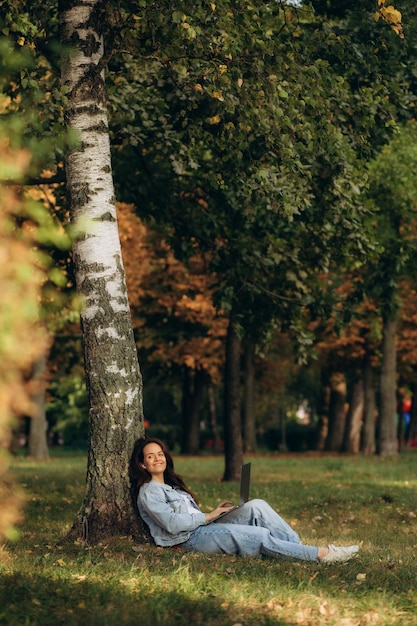 Portrait of pretty young woman sitting on green grass in park with legs crossed during summer day while using laptop and wireless earphone for video call