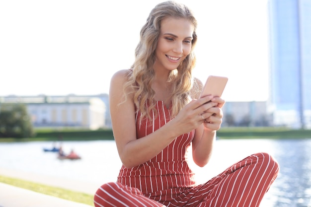 Portrait of pretty young woman sitting on riverbank with legs crossed during summer day, using smartphone.