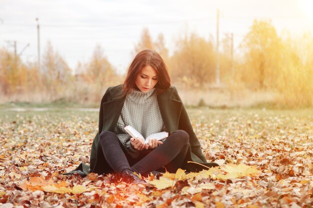 Portrait of pretty young woman of Slavic appearance in casual clothing in autumn, reading book in clearing. Cute model walking in Park in golden fall against background of nature. Copy space