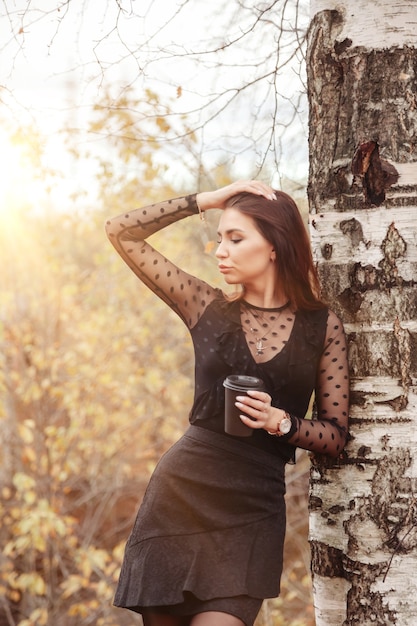 Portrait of pretty young woman of Slavic appearance in dark dress with cup of coffee in autumn, standing next to birch tree against background of an autumn Park. Golden fall. Copy space