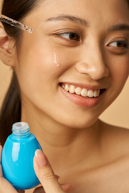 Portrait of pretty young woman smiling aside, dropping oil serum with pipette on face for fresh perfect skin isolated over beige background. Beauty treatment concept