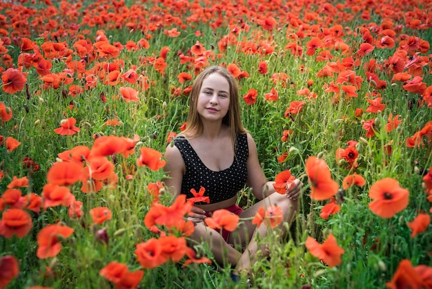 Ritratto di una donna abbastanza giovane in abiti sportivi che si siede nel campo di papaveri fioriti. goditi la libertà in una giornata estiva