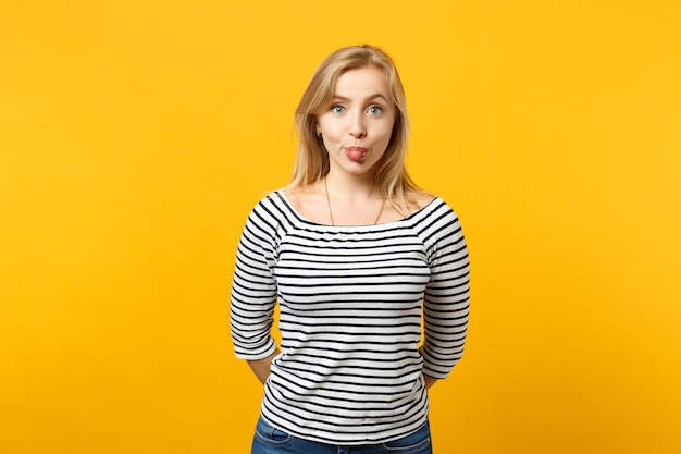 Portrait of pretty young woman in striped clothes looking camera, showing tongue isolated on yellow orange wall background in studio. People sincere emotions, lifestyle concept. Mock up copy space.