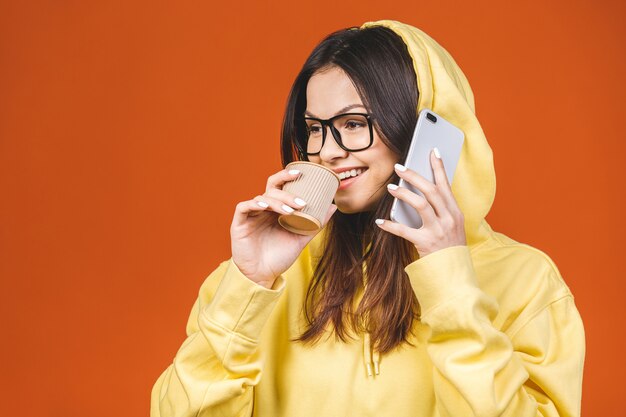 Portrait of a pretty young woman using mobile phone while holding takeaway coffee cup isolated over orange background.