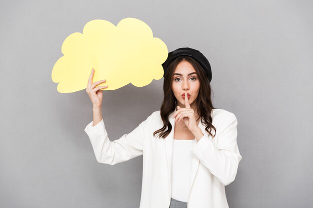 Portrait of a pretty young woman wearing beret standing isolated over gray background, showing empty speech bubble, showing silence gesture