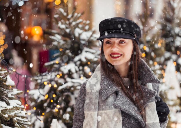Портрет красивой молодой женщины в стильной черной шляпе перед Рождеством на фоне города в снежное время