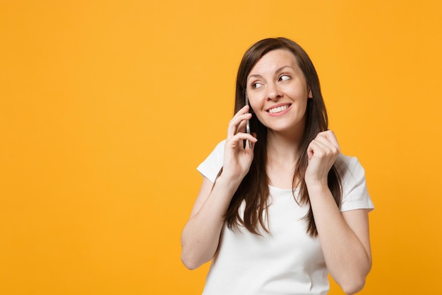 Ritratto di bella giovane donna in abiti casual bianchi che guarda da parte, parlando al telefono cellulare isolato su sfondo giallo brillante parete arancione in studio. concetto di stile di vita delle persone. mock up spazio di copia.