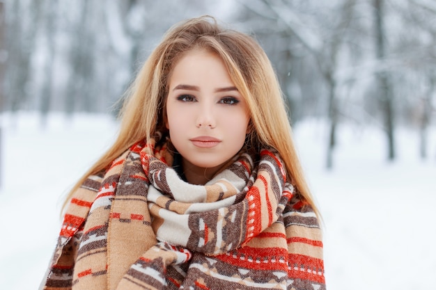 Portrait of a pretty young woman with brown eyes with beautiful makeup  with long blond hair in a woolen vintage warm scarf in a snowy park. Cute girl on a walk.