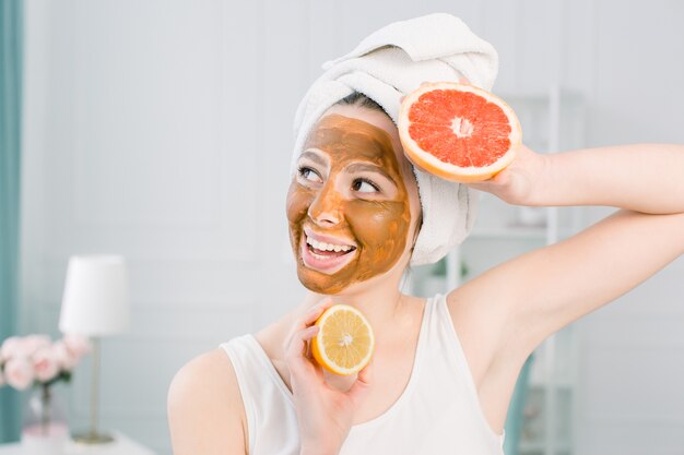 Portrait of pretty young woman with clay mask on her face holding slices of lemon and grapefruit, facial care