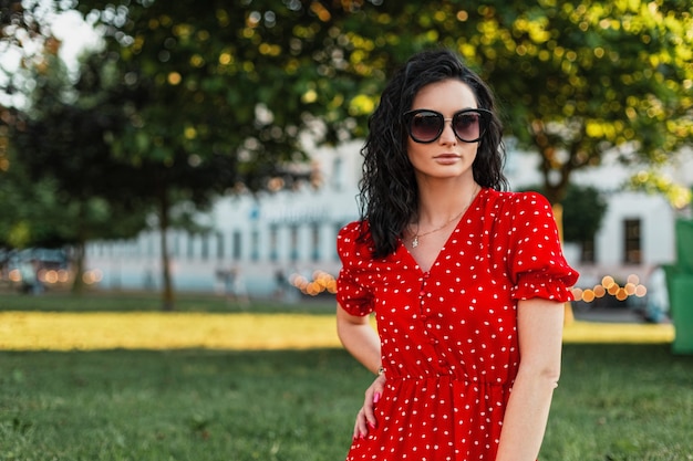 Portrait of a pretty young woman with curly hair in fashionable sunglasses and a red dress walks on the street