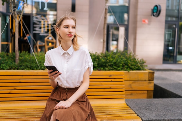 Portrait of pretty young woman woman sitting on yellow stone bench holding smartphone in hands and smiling looking away on modern city street