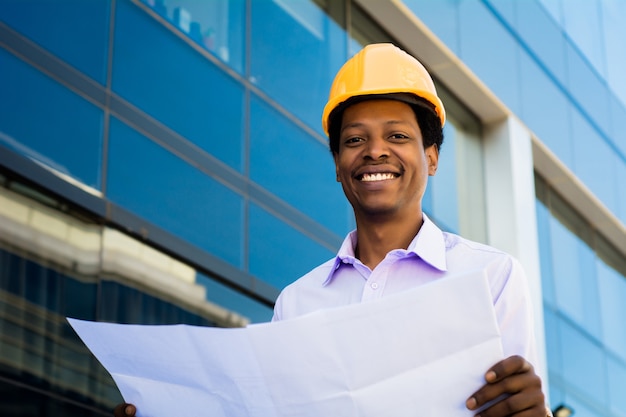 Portrait of professional architect in helmet looking at blue prints outside modern building. Engineer and architect concept.