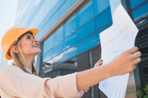 Portrait of professional architect wearing yellow helmet and looking at blue prints outside modern building. Engineer and architect concept.