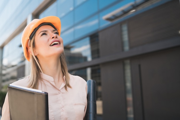 Portrait of professional architect woman wearing yellow helmet and looking at modern building outdoors. Engineer and architect concept.