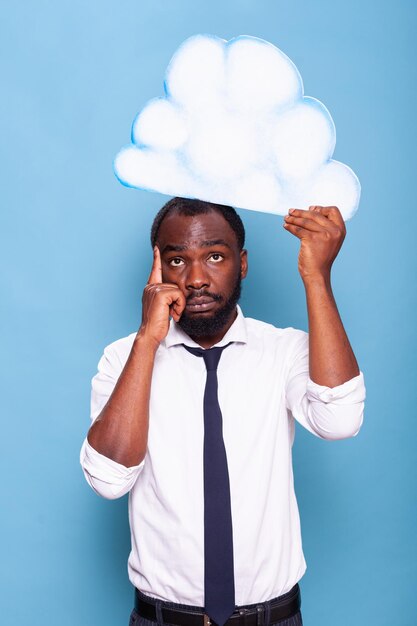 Portrait of professional businessman pointing at white paper idea cloud standing in front of blue background. African american entrepreneur thinking under thought bubble.