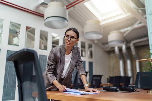 Portrait of professional businesswoman in glasses standing in modern office with documents