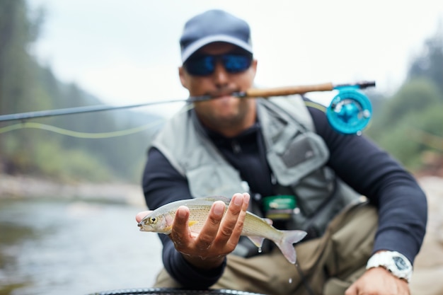 Portrait of professional fisherman showing rainbow trout in mountain river. Happy man holding his catch during favorite hobby on fresh air.