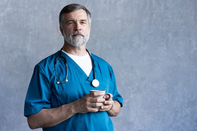 灰色の背景に立ってカメラを見ながら、カップを保持しているプロの成熟した外科医の肖像画。