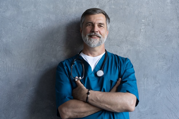 회색 배경에 서 있는 동안 카메라를 보고 있는 전문 성숙한 외과 의사의 초상화.