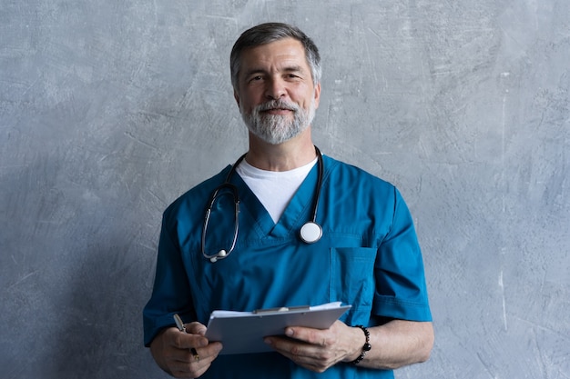 Portrait of professional mature surgeon looking at camera while standing against the grey background.