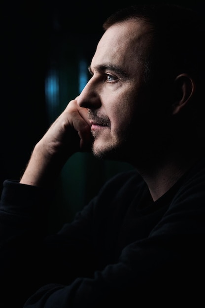 Photo portrait in profile of a pensive guy in a black jacket in a dark room who leaned on his hand blue eyes