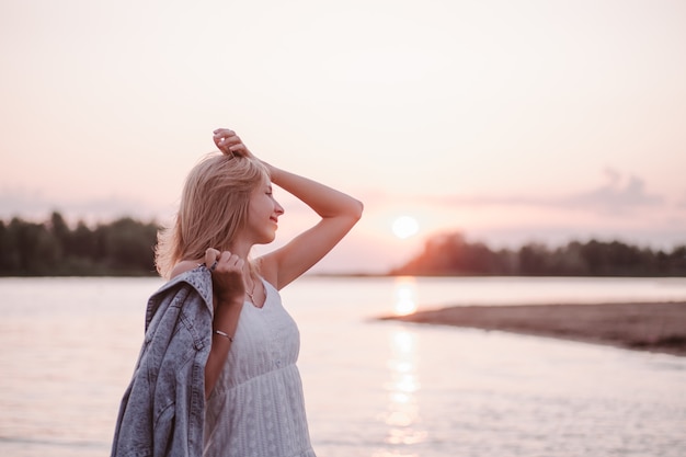 Portrait in profile of a young woman on the beach a beautiful blonde in a white summer dress stands ...