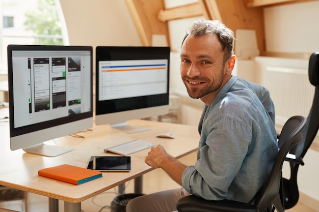 Portrait of programmer smiling at camera while sitting at his workplace with computer monitors and working with software