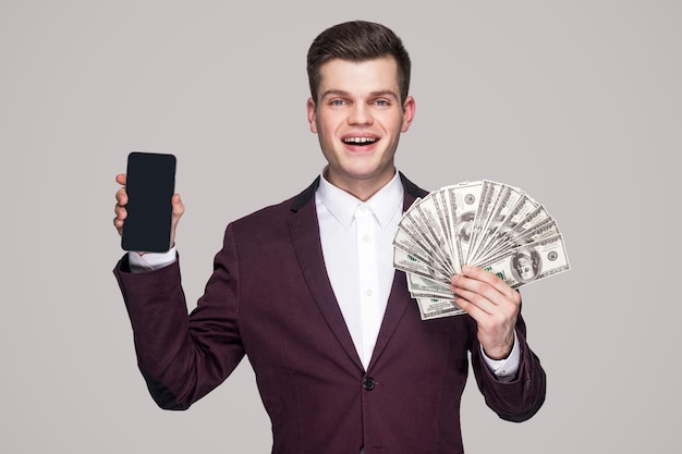 Portrait of prosperous atractive young businessman in classic violet coat standing, looking at camera and holding fan of money and smart phone display. Indoor studio shot, isolated on gray background