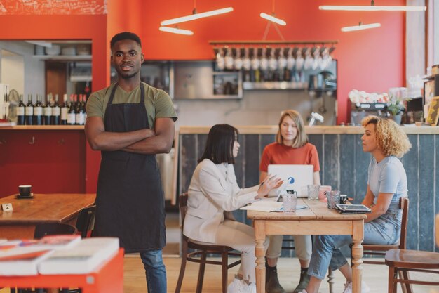 Portrait of a proud African American man in his cafe restaurant multiethnic women in background sitting on table working together