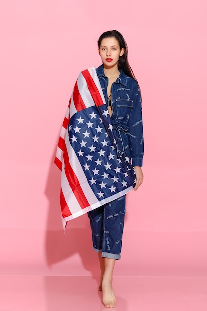 Portrait of proud female athlete wrapped in American Flag against pink background. Muscular young woman looking confidently at camera. female protesting for equality and women empowerment