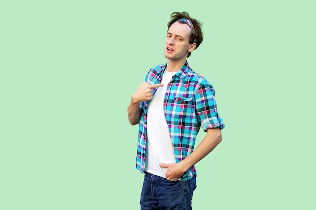 Portrait of proud young man in casual blue checkered shirt and headband standing, showing and pointing himself and looking at camera with pride. indoor studio shot, isolated on light green background.