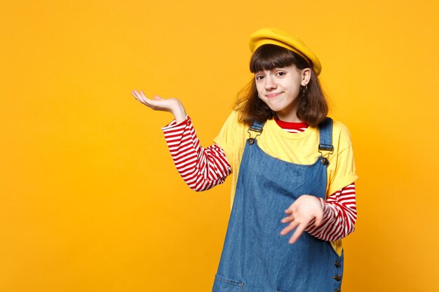 Portrait of puzzled girl teenager in french beret, denim sundress spreading pointing hand aside isolated on yellow background in studio. people sincere emotions, lifestyle concept. mock up copy space