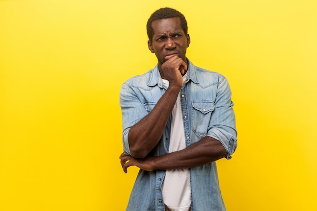 Portrait of puzzled pensive man in denim casual shirt with rolled up sleeves looking up and thinking intensely with doubting uncertain expression indoor studio shot isolated on yellow background