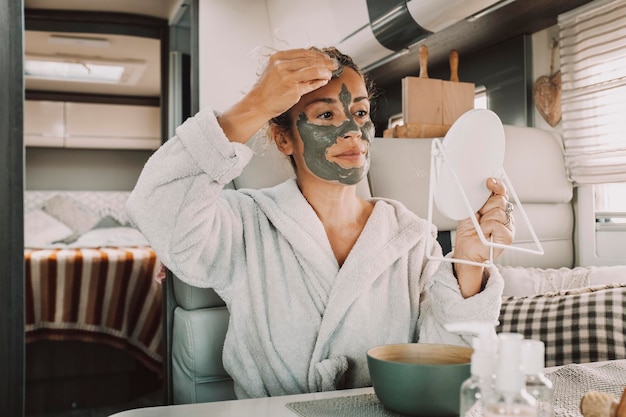 Portrait of real adult woman applying green mud beauty mask on face using mirror and hands inside a camper van Alternative lifestyle freedom people travel and nature Daily routine young female alone