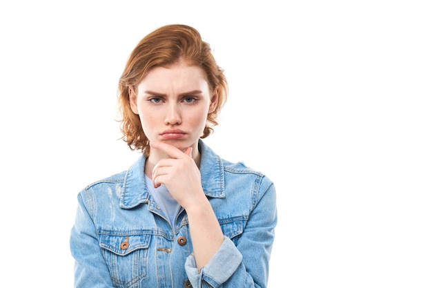 Portrait of red haired dreamy lovely lady, youth. Redhead woman in blue jeans displeased thinks, chooses, decides, dilemmas, isolated white background. Girl student is unhappy