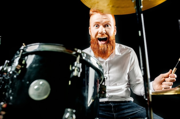 Portrait of a red-haired emotional man playing drums and cymbals and holding a stick