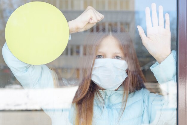 Ritratto di una ragazza dai capelli rossi attraverso la finestra in una maschera protettiva come concetto di quarantena e isolamento dovuto al coronavirus covido-19, alla chiusura del confine e alla disunità sociale nella pandemia