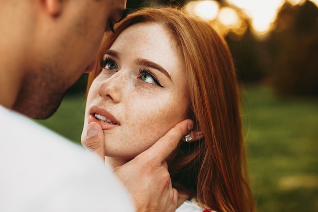 Портрет рыжеволосой женщины с веснушками и зелеными глазами, смотрящей на своего парня, пока он трогает ее губы пальцем на фоне заката во время свидания
