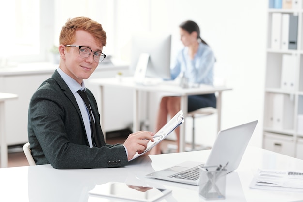 Portrait of red headed young businessman smiling while sitting at desk in office, copy space